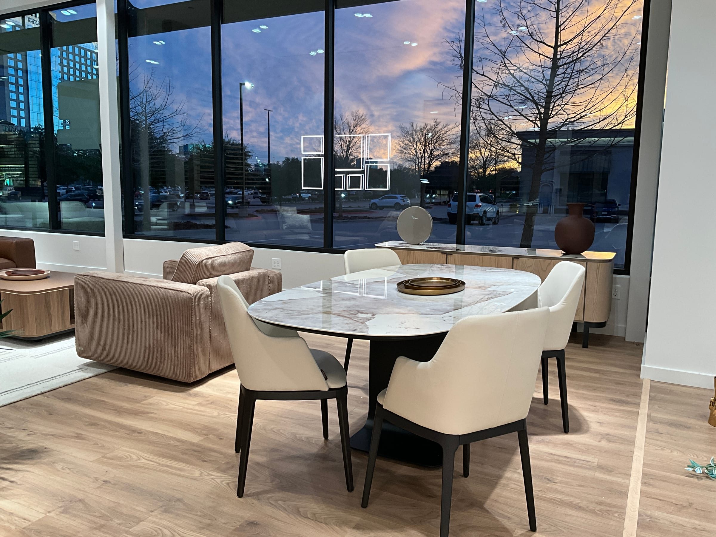 Dining vignette with a round marble-top table and white dining chairs, evening skyline through floor-to-ceiling windows.