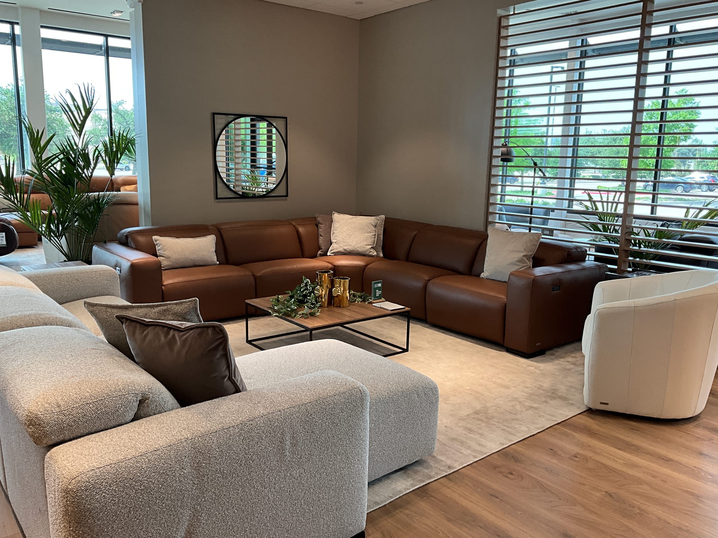 Cognac leather sectional staged in a corner beneath a round mirror, near wood-slat window shades.